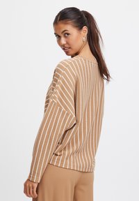 Pull en maille marron avec des rayures blanches horizontales, épaules tombantes et coupe décontractée, associé à un pantalon marron clair.