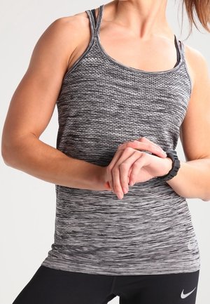 Kvinde i gråt mønstret atletiktop og sorte leggings justerer et sort smartwatch på sit håndled mod en ensfarvet baggrund.