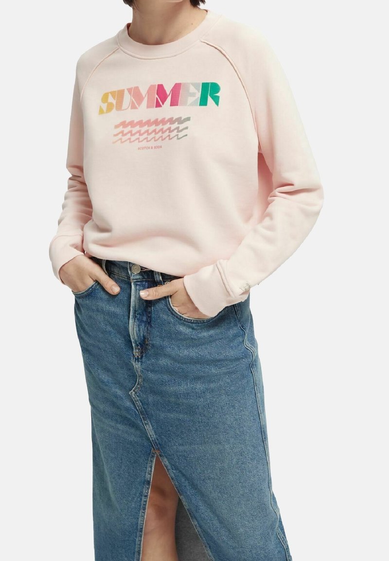 Felpa rosa chiaro con la scritta "SUMMER" in testo colorato e motivi geometrici. Abbinata a una gonna in denim blu con uno spacco frontale.