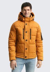 Orangenes Puffer-Jackett mit Kapuze, ausgestattet mit schwarzen Reißverschlusstaschen, Knopfverschlüssen und gestepptem Design. Getragen über einem schwarzen Shirt.