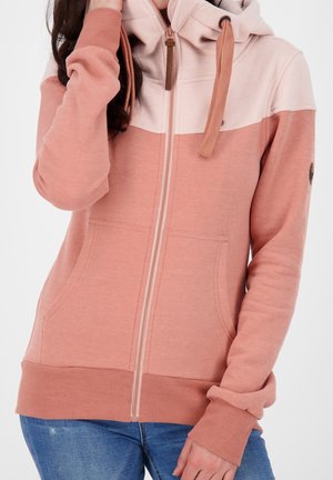 Vrouw draagt een tweekleurige roze zip-up hoodie met voorzakken en trekkoorden, gecombineerd met blauwe spijkerbroek, tegen een effen achtergrond.