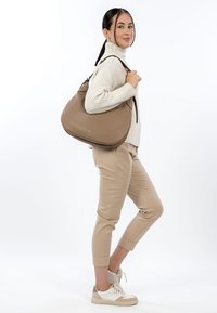 Pruun nahast hobo-kott, sileda tekstuuri, kumerate vormide ja kahe käepidemega. Modell kannab helehalli kampsunit ja beeže pükse, seisab vabas poosis.