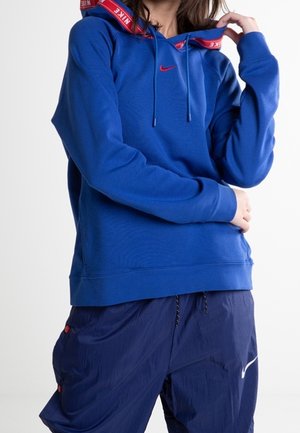 Osoba w niebieskiej bluzie z kapturem Nike z czerwonym logo i pasujących niebieskich spodniach dresowych Nike, stojąca ze skrzyżowanymi rękami i jedną ręką przy szyi.