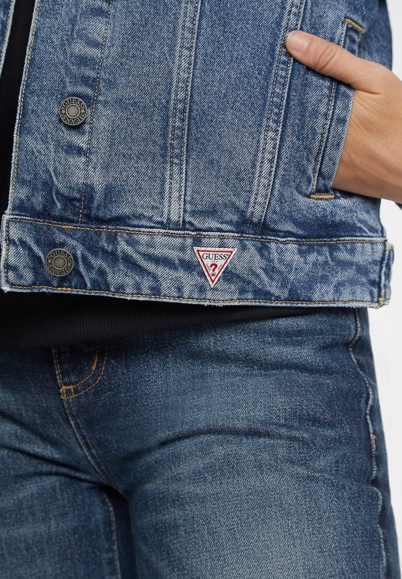 Chaqueta de mezclilla en azul claro con un acabado texturizado, que presenta un parche en forma de triángulo con "GUESS", botones de metal y dos bolsillos laterales inferiores.