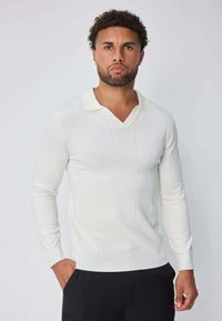 Homme avec cheveux bouclés et barbe, portant un pull blanc à col en V à manches longues et un pantalon noir, debout avec une main dans la poche.