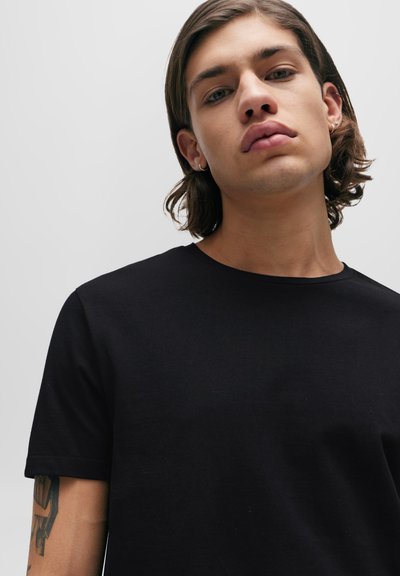 T-shirt en coton noir avec un col rond, des manches courtes et une texture lisse. Un tatouage est visible sur l'avant-bras gauche.