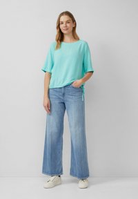 Türkises kurzärmliges Oberteil mit seitlichem Knoten, kombiniert mit hellblauen weit geschnittenen Jeans und weißen Turnschuhen. Einfarbige Farben, glatter Stoff.