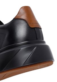 Tomaia nera della scarpa sportiva con suola nera sollevata, linguetta sul tallone in pelle marrone e suola esterna in gomma marrone testurizzata visibile.