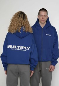 Felpa blu con zip e grande logo bianco "MULTIPLY" sul retro. Dotata di cappuccio e tasche frontali; abbinata a pantaloni grigi.