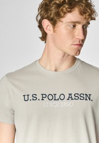T-shirt a maniche corte grigio chiaro in cotone. Presenta sulla parte anteriore il testo ricamato blu navy "U.S. POLO ASSN." e "SINCE 1890".