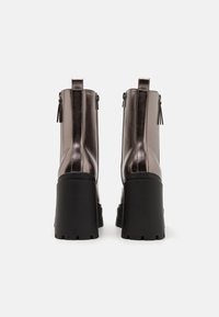 Botas metálicas hasta el tobillo en bronce oscuro con un alto y grueso tacón negro. Cuenta con un cierre de cremallera y borlas decorativas en la parte trasera.
