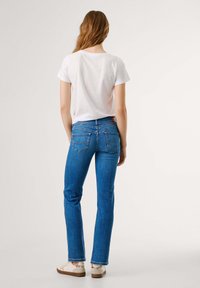 Kvinne med langt hår iført hvit t-skjorte, blå rette jeans og hvite sneakers, som står med ryggen til på en ensfarget bakgrunn.