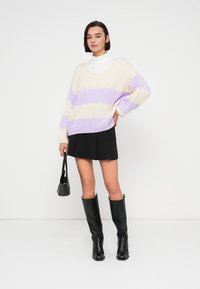 Pull surdimensionné en maille crème à rayures violettes, associé à une jupe plissée noire et des bottes noires brillantes montantes jusqu'aux genoux. Sac à main noir.