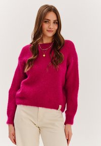 Różowy sweter z dzianiny z długimi rękawami, okrągłym dekoltem i teksturą przypominającą miękką, puszystą przędzę. W połączeniu z jasnymi spodniami.