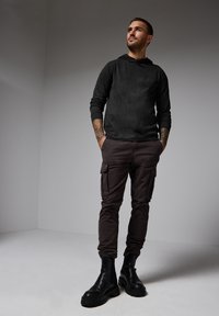 Svart huvtröja med långa ärmar, kombinerad med mörkbruna cargobyxor med sidofickor och svarta ankelkängor. Slät textur.