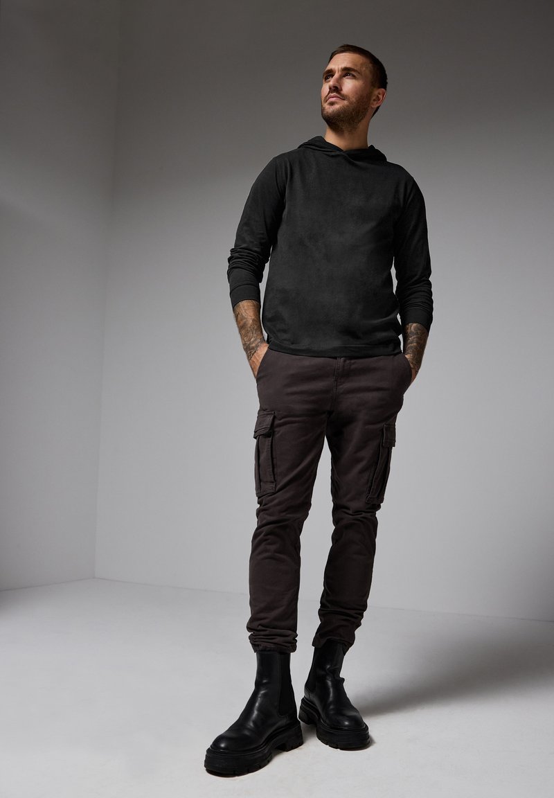 Svart huvtröja med långa ärmar, kombinerad med mörkbruna cargobyxor med sidofickor och svarta ankelkängor. Slät textur.
