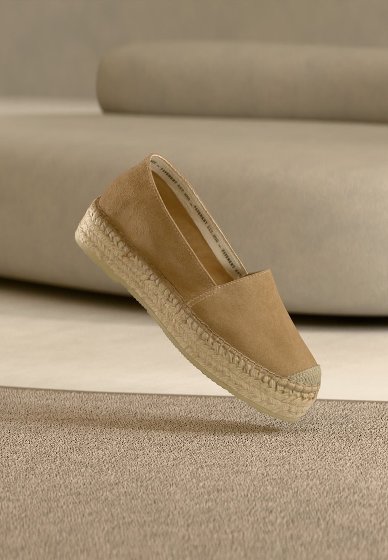Espadrille en suède marron clair avec semelle en jute tissée flottant au-dessus d'un tapis texturé beige et d'un fond de canapé beige doux.