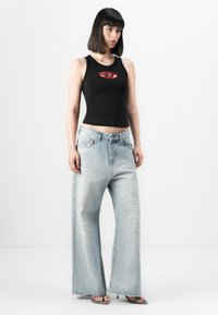 Femme avec des cheveux raides et foncés portant un haut sans manches noir avec un logo, un jean large déchiré bleu clair et des sandales à talons ouvertes.
