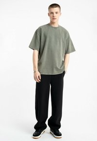 Przestronna oliwkowa koszulka z bawełny z okrągłym dekoltem i krótkimi rękawami, w połączeniu z luźnymi czarnymi spodniami i czarnymi butami.