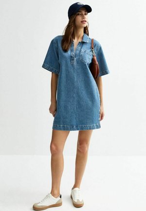 Denim polo jurk in lichtblauw, korte mouwen, kraag en een borstzakje. Gecombineerd met witte sneakers en een marineblauwe pet met bruine accenten.