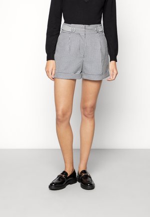 Kvinde iført sort langærmet top, sort-hvide ternede shorts med højtaljede knapper og sorte loafers med metaldetalje.