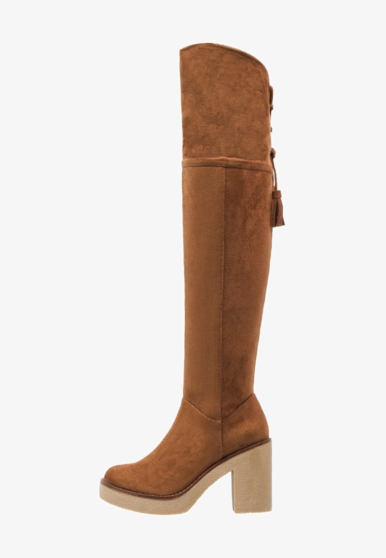 Bottes en daim marron au-dessus du genou avec un talon plateforme épais, un bout rond et des franges décoratives à l'arrière. Texture lisse et design ajusté.