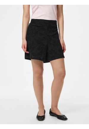 Kvinne iført svarte broderte shorts, blekrosa ermeløs topp og svarte ballettsko, stående på hvit bakgrunn.