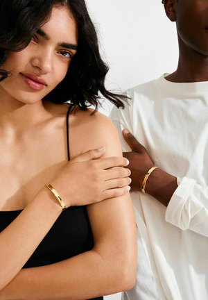 Gouden armbanden gedragen op blote armen, met een textuur en gepolijst ontwerp. Eén armband aan een lichtgekleurde arm, een andere aan een donkergekleurde arm.