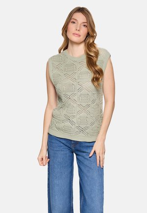 Mujer con cabello largo y ondulado, vistiendo un top de punto sin mangas color verde claro con un patrón geométrico y jeans de mezclilla azul, de pie frente a un fondo blanco.