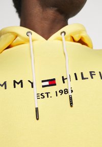 Sudadera amarilla de algodón, con capucha ajustable, logotipo en relieve "TOMMY HILFIGER" con detalle de bandera en rojo, blanco y azul, establecida en 1985.