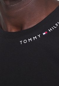 Mustapaita, jossa on pyöreä pääntie ja valkoista "TOMMY HILFIGER" -tekstiä logovihjeillä kauluksen lähellä. Sujuva pinta.