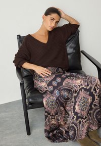 Pull en maille côtelée marron avec col en V et manches longues. Jupe noire fluide avec un motif cachemire en tons roses et bleus. Chaise en cuir noir.