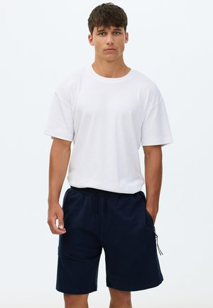 Joven con cabello corto y oscuro, vistiendo una camiseta blanca holgada y pantalones cortos azul marino oscuros hasta la rodilla, de pie contra un fondo claro y sencillo.
