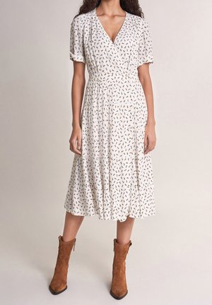 Kobieta ubrana w białą sukienkę midi z drobnym brązowym kwiatowym wzorem, krótkimi bufkami, dekoltem w serek oraz brązowymi zamszowymi botkami, stojąca na beżowym tle.