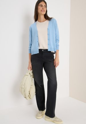 Lichtblauwe cardigan met knopen, beige top, zwarte jeans, houdt een crèmekleurige gequilt tas vast en draagt beige sneakers.