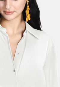 Blusa bianca in satin con colletto classico, chiusura frontale con bottoni e texture liscia, abbinata a orecchini gialli a balze.