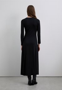 Robe noire à manches longues, ajustée, avec un ourlet légèrement évasé. Texture douce, design minimaliste, et détail de boutons dans le dos. Portée avec des bottes montantes jusqu'aux genoux.