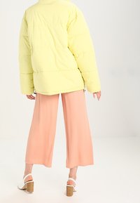 Chaqueta amarilla acolchada con un cuello alto, combinada con pantalones rosa de pierna ancha. El conjunto incluye sandalias de tacón blancas. Texturas suaves visibles.