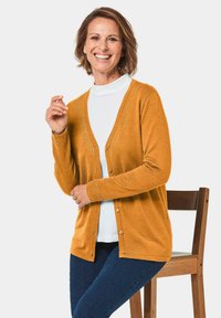 Senfgelber Cardigan mit V-Ausschnitt, Knopfverschluss und dekorativen Nähten entlang des Ausschnitts und der Bündchen. Kombiniert mit einem weißen Oberteil und blauen Jeans.