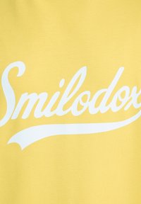 Geel katoen stof met grote witte tekst die leest "Smilodox," met een gebogen accentlijn onder de tekst.