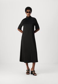 Rochie neagră, până la gleznă, cu guler înalt și mâneci scurte. Textură netedă. Poartă sandale negre cu toc, având accente aurii.