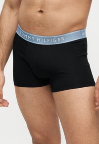 Άνδρας κορμός με μαύρα boxer Tommy Hilfiger με ανοιχτό μπλε μέση, δείχνοντας μέρος των χεριών και των άνω ποδιών.