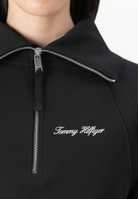 Black zip-up top made of ribbed fabric, featuring a high collar and a silver zipper. "Tommy Hilfiger" is embroidered in white.