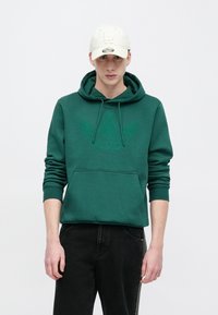 Πράσινο φούτερ με κουκούλα και μεγάλο λογότυπο σε τονικούς χρωματισμούς στο μπροστινό μέρος, τσέπη τύπου καγκουρό στο μπροστινό μέρος και ριμπ μανσέτες, συνδυασμένο με μαύρο παντελόνι και μπεζ καπέλο.