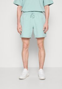 Vaaleanvihreät puuvillashortsit, joissa on joustava vyötärö, nyöri ja sivutaskut, yhdistettynä valkoisiin tennareihin. Yksinkertainen, urheilullinen muotoilu.