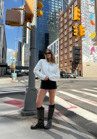 Fehér hosszú ujjú felső húzózsinórral, fekete tűsarkú miniszoknya és magas fekete csizmák városi utcakép hátterében.