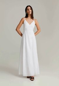 Vestido largo sin mangas de color blanco con escote en V, corsé ajustado y falda fluida. Textura suave, diseño minimalista y sandalias negras con tira al tobillo.