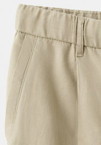 Pantalon beige avec taille élastique, poche latérale et passants pour ceinture, en tissu texturé.