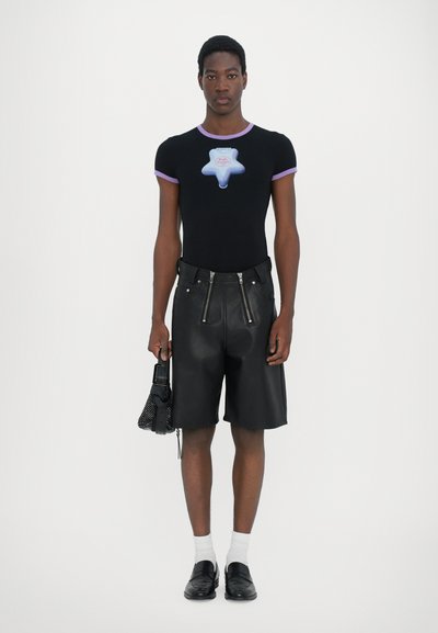 Schwarzes, eng anliegendes T-Shirt mit lila Ausschnitt, das mit einem blauen Stern-Graphic kombiniert wird, sowie hochtaillierten schwarzen Leder-Shorts und schwarzen Slippern.