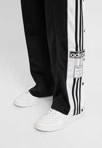 Svart och vitt träningsbyxor med tryckknappar, tre ränder längs sidorna och Adidas-logga på låret. Ett par vita sneakers.